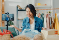 Young Asia lady fashion designer using mobile phone receiving purchase order and showing clothes recording video live streaming online at shop. Small business owner, online market delivery concept.