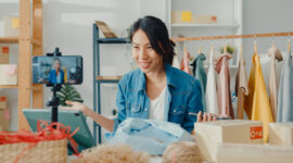 Young Asia lady fashion designer using mobile phone receiving purchase order and showing clothes recording video live streaming online at shop. Small business owner, online market delivery concept.
