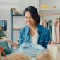 Young Asia lady fashion designer using mobile phone receiving purchase order and showing clothes recording video live streaming online at shop. Small business owner, online market delivery concept.